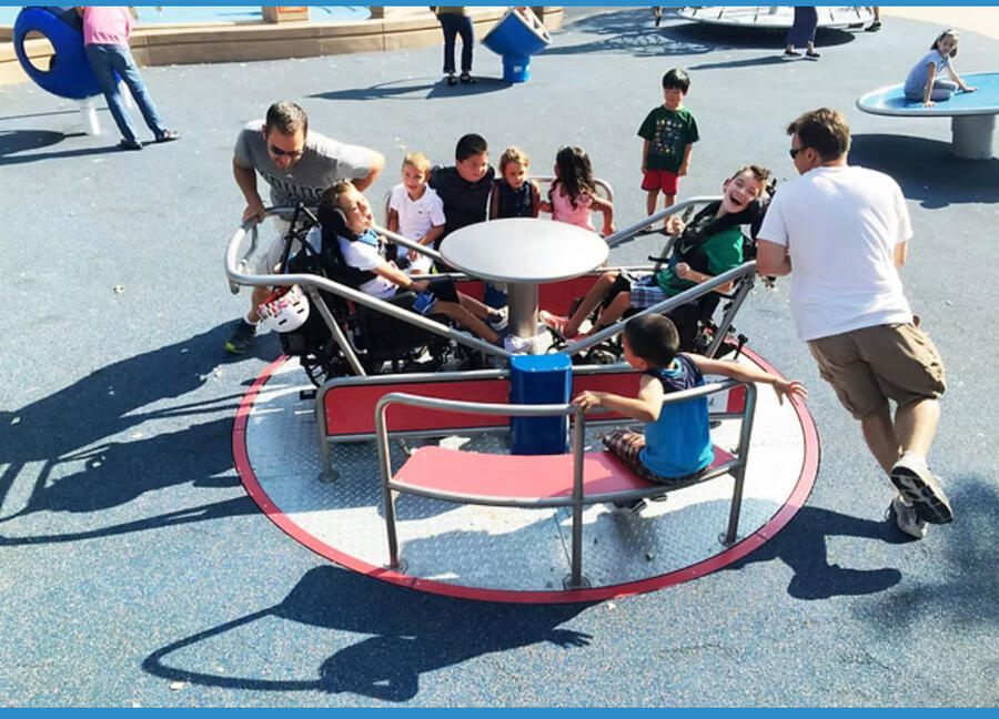 @magicalbridgefoundation alt= Fathers pushing happy children around a merry go round in a park