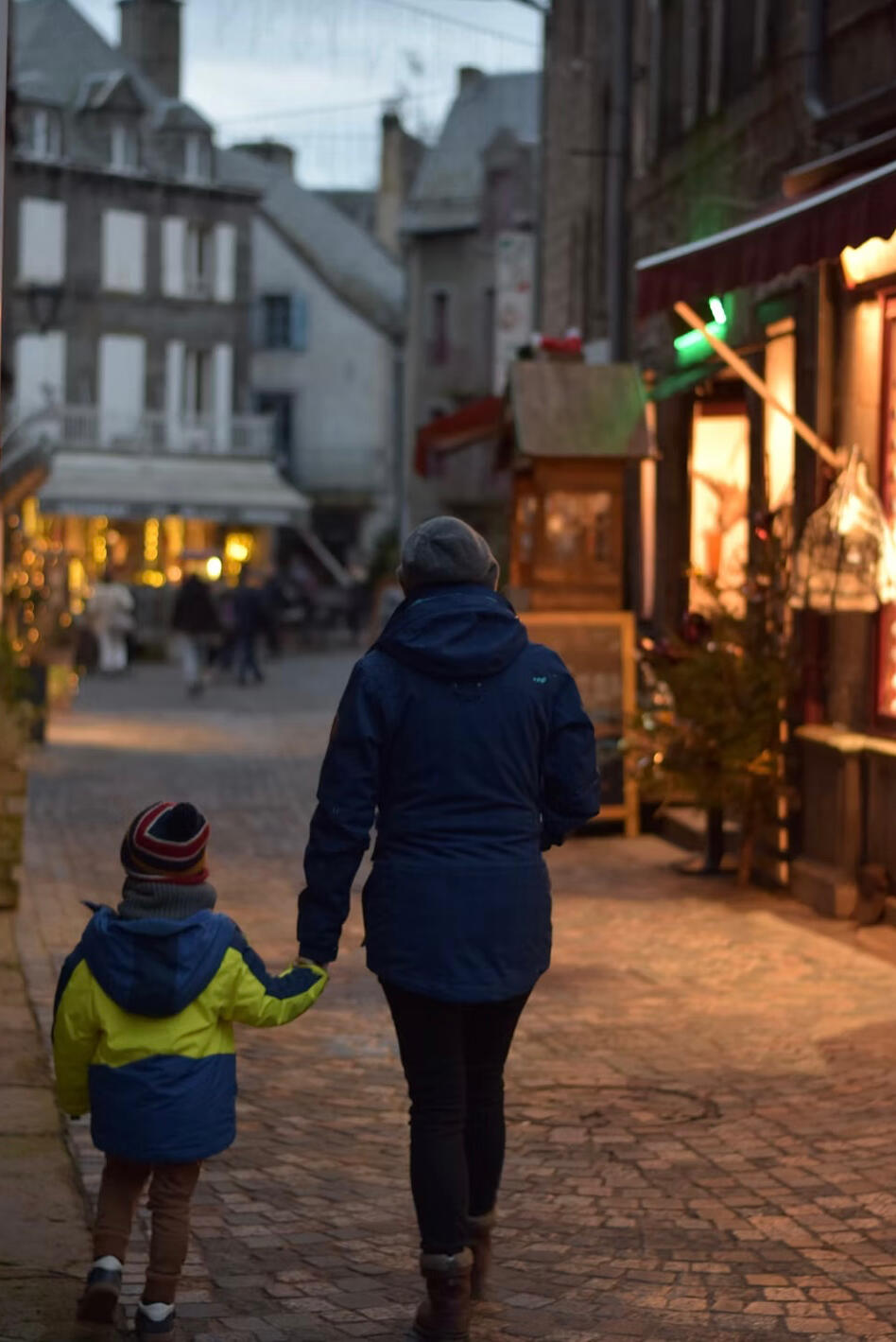 Parent and young child wearing winter clothes walking away from the camera on a quaint cobblestone street at dusk with shop windows glowing with a golden light