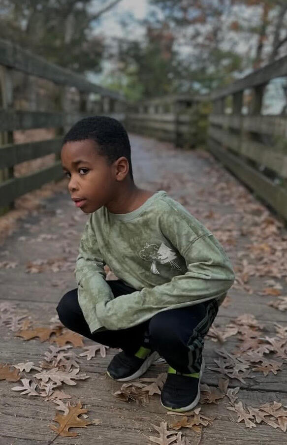A young black boy wearing a sweatshirt and jeans poses for a photo in the middle of a park footbridge in the fall.
