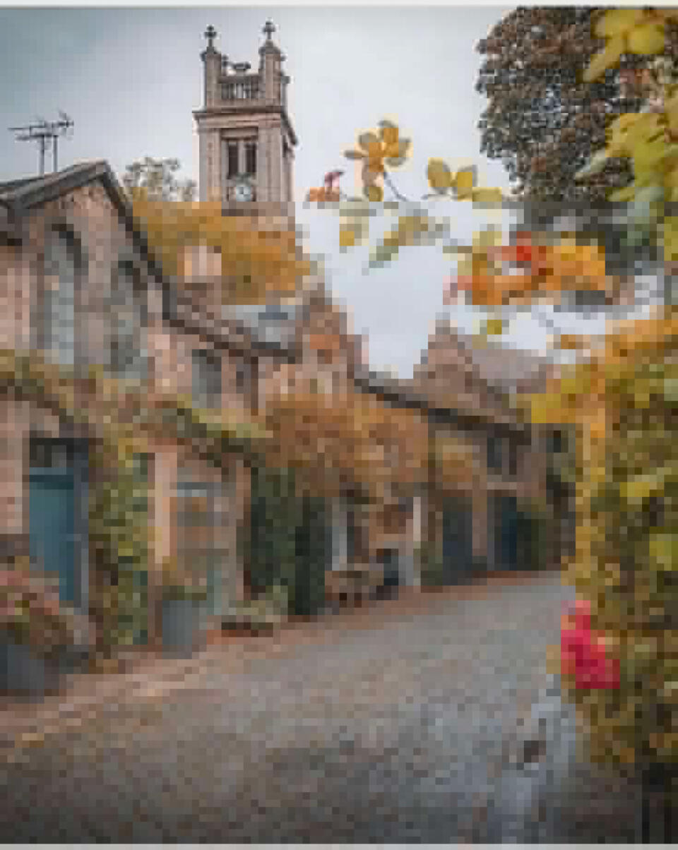European village with stone and timber homes and shops on a curving cobblestone street in early spring with a few colorful flowers in broad daylight. The camera is focused on the bell and clock tower of what appears to be a greystone Catholic cathedral.