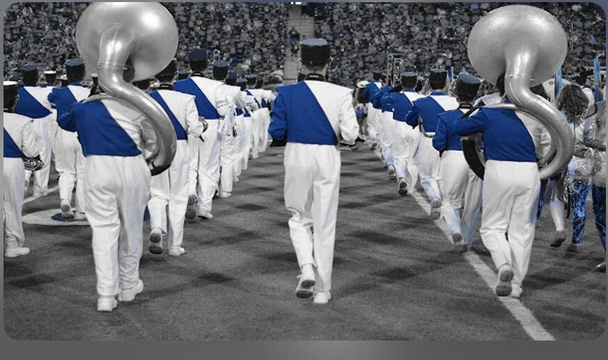 A high school marching band wearing white pants and blue uniform tops march away from the camera.