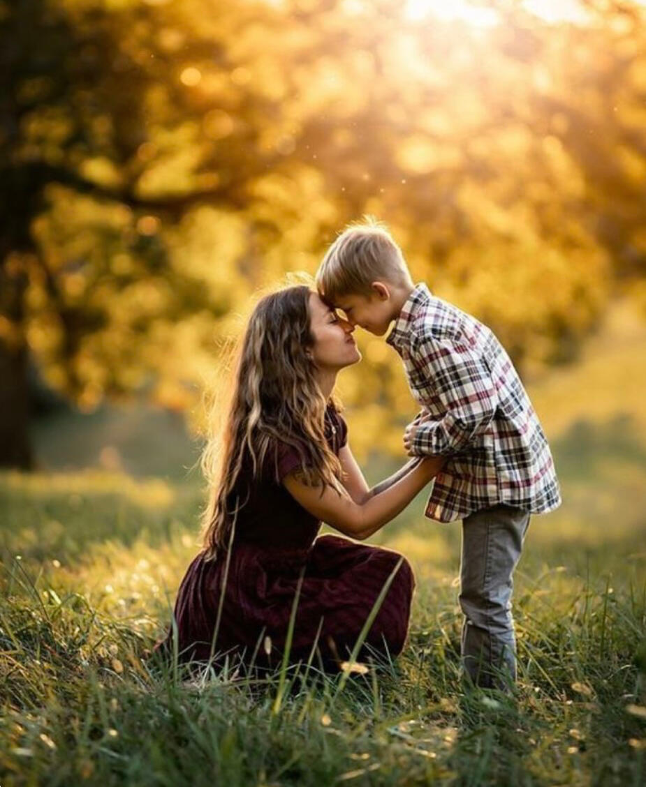 Mother and son in the center of a meadow surrounded by yellow and gold leafy trees in filtered sunlight. Both mother and son are smiling, holding hands, and touching foreheads. The mother is crouched down to be at her son’s height.