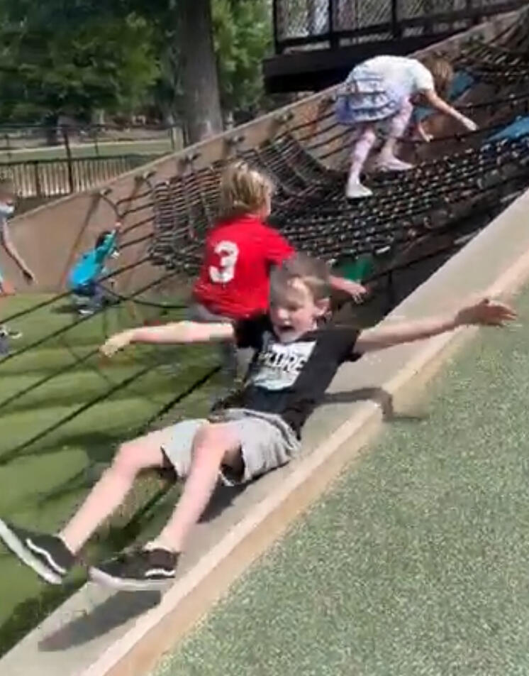 3 children at a playground. Two are climbing up a small hill. The boy in the foreground is sliding down a slide with arms and legs spread wide.