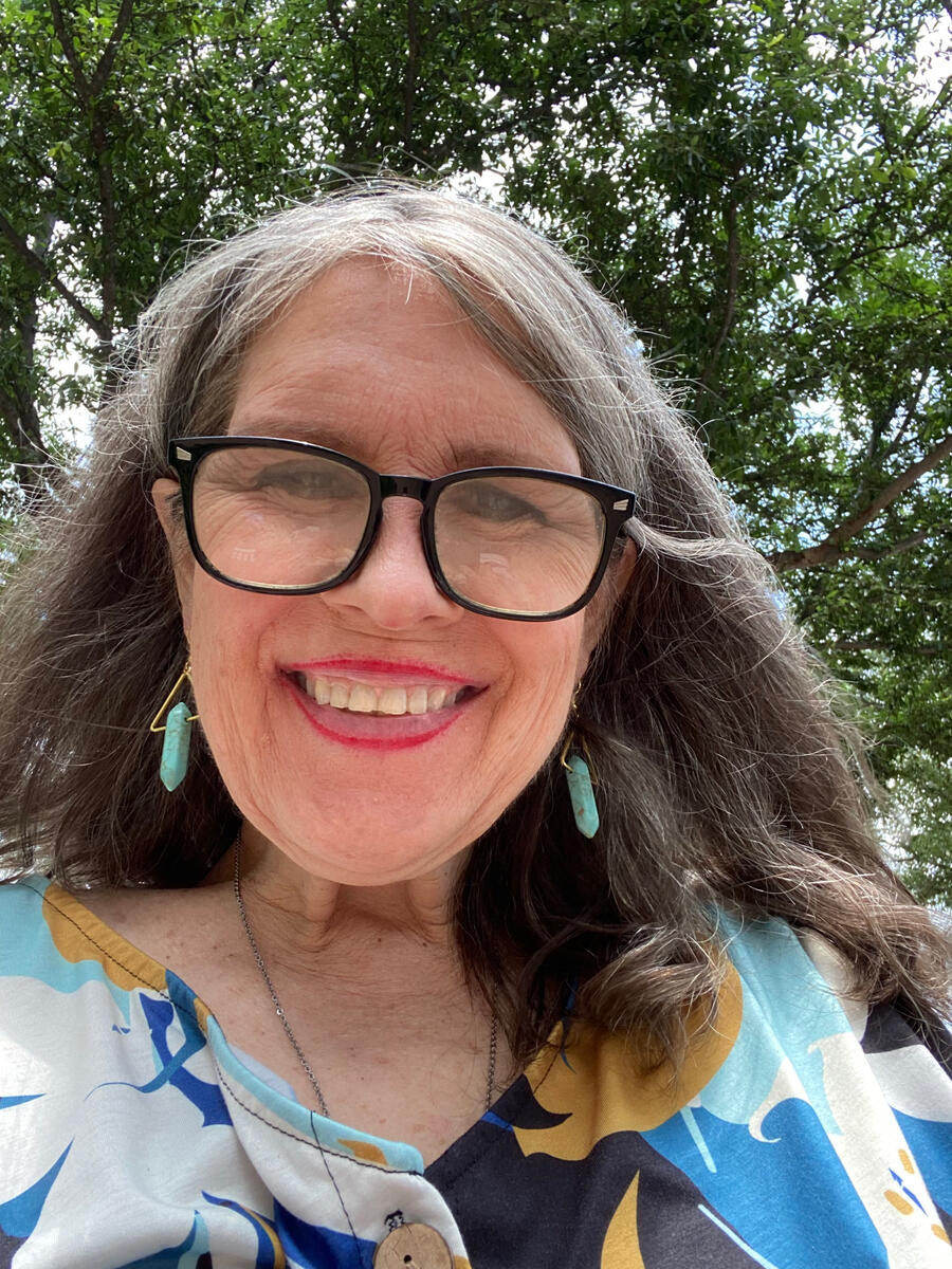 alt= Image of advocate Donna Earnest. She is in her 60’s, long brown hair, big black nerdy glasses, smiling widely and wearing a turquoise print dress.