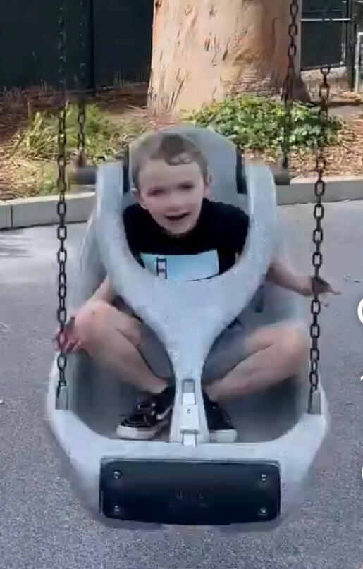alt= White little boy with short hair around 6 years old sitting cross legged on a large plastic swindle smiling in mid-swing