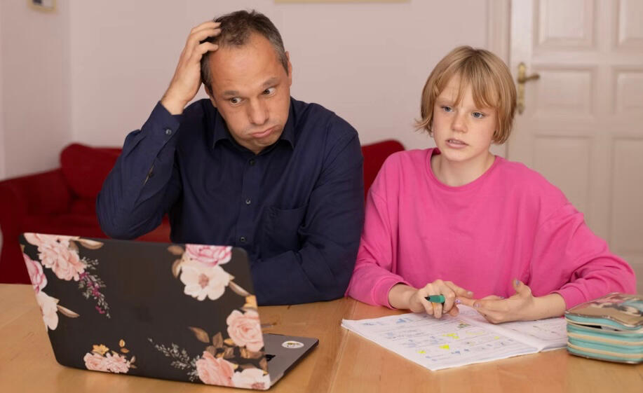 alt= Father trying to help his daughter with homework. Dad is wearing a black sweater, scratching his head, and blowing out air from his cheeks as he stares at a laptop. The daughter is wearing a bubblegum pink sweater and has short blonde hair. She is loo